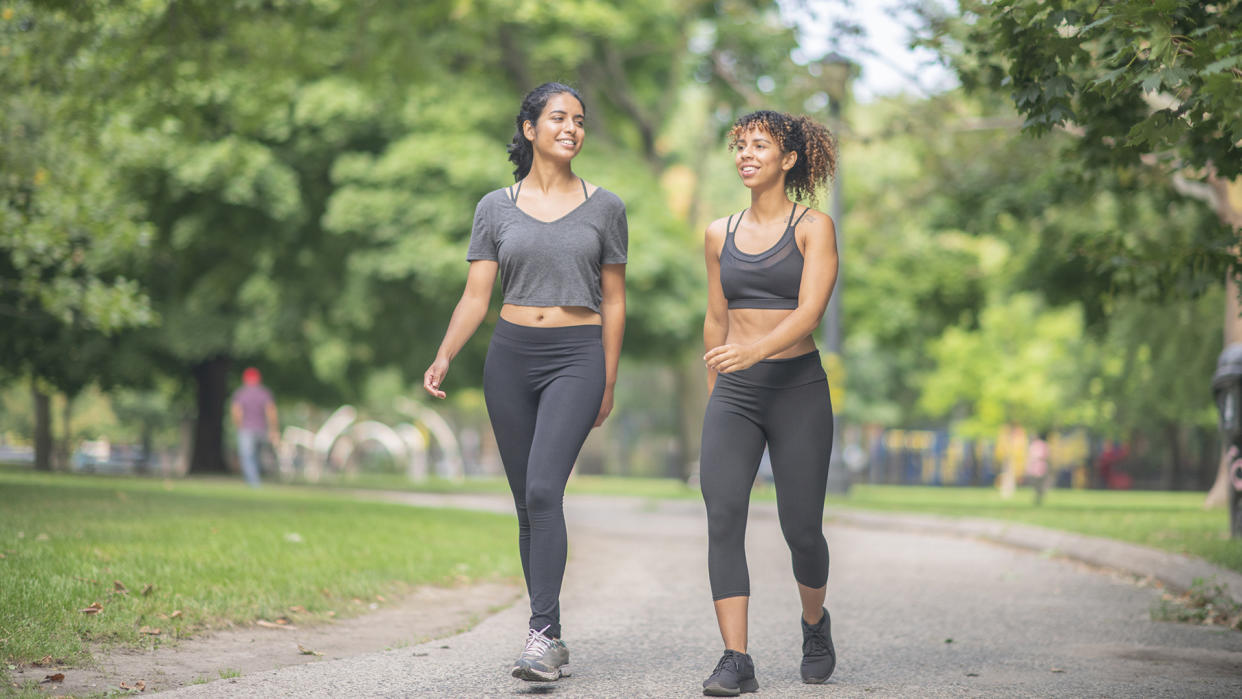  A pair of diverse friends smile as they workout in the park together. They are walking during the afternoon. 