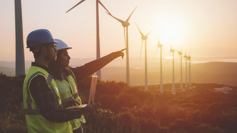 Young electrical engineer woman and business man standing in front of wind turbines checking and working about technical problems and writes the results of measurements with laptop pc in wind power plant electric energy station.