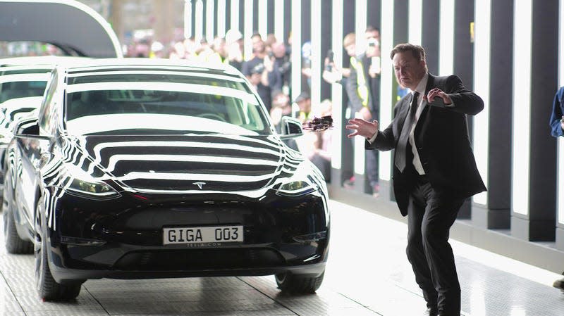 Elon Musk waving his hands and making a face in front of a Tesla car.