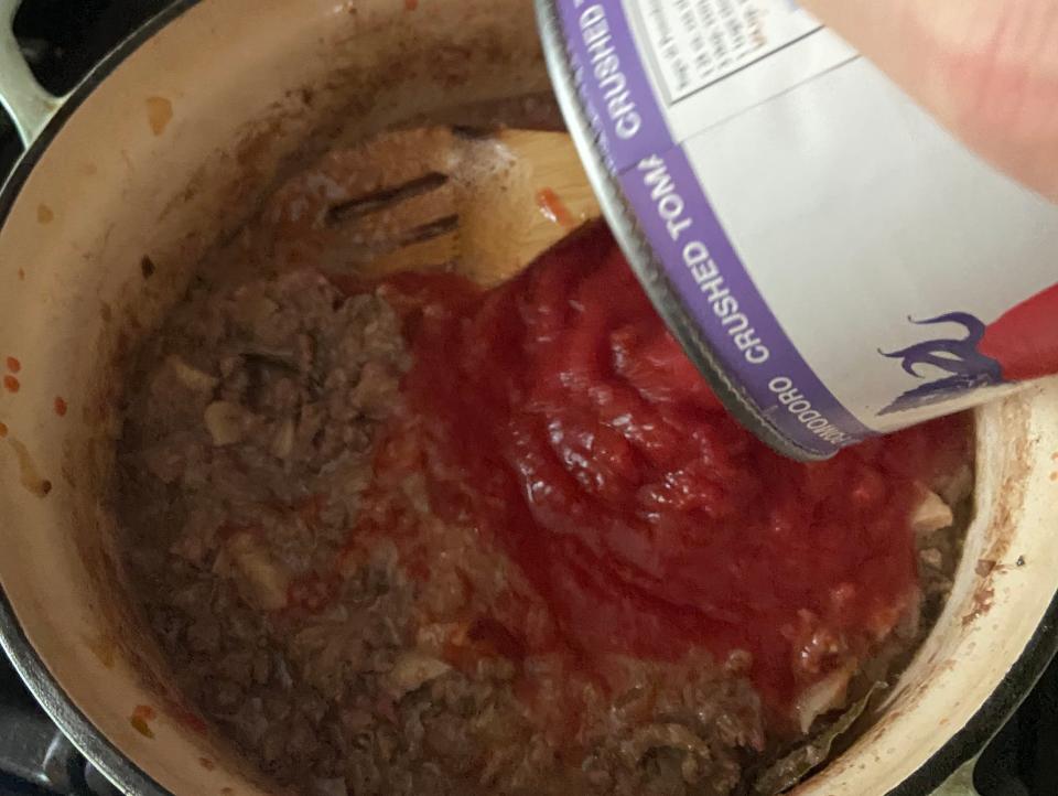 hand pouring can of crushed tomatoes into a pot of meat sauce on the stove