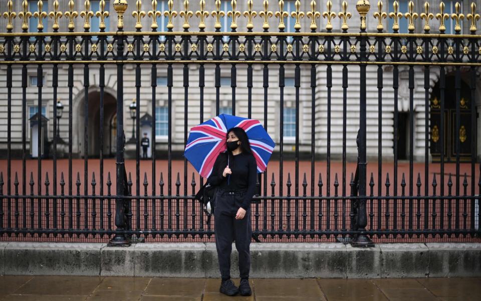 A woman wearing a protective face covering, shelters from the rain under a Union flag umbrella outside Buckingham Palace in central London -  DANIEL LEAL-OLIVAS/AFP