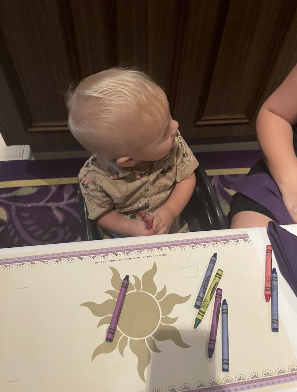 A toddler with light hair sits at a table with crayons and a sun-themed mat, looking to the side. An adult's arm is partially visible next to the child