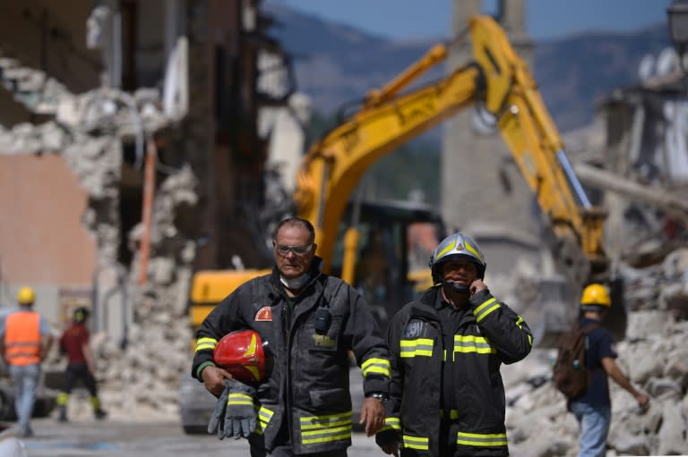 Firefighters walk past an excavator used to search for victims in the remains of a building in the central Italian village of Amatrice on August 25, 2016, a day after a 6.2-magnitude earthquake struck the region