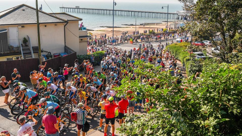 A large group of cyclists wearing helmets and cycling gear in Felixstowe, with spectators taking photographs and cheering them on.