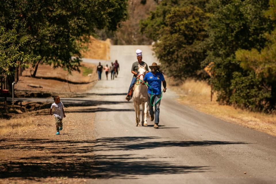 A child running to keep up with a man leading a woman on a horse along on a dirt road, as others walk in the distance