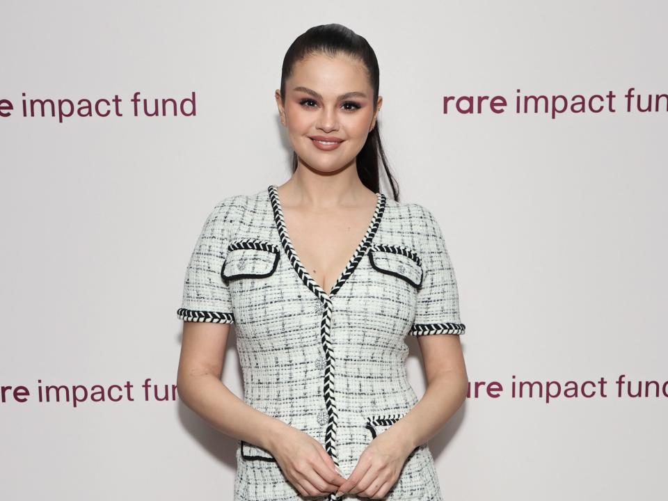 selena gomez in a black and white dress and black tights. the dress sits at mid-thigh and has a v neckline, as well as pockets on both breasts and near her waist. gomez is smiling widely, with her hair pulled back into a ponytail. text on the wall behind her reads "rare impact fun"