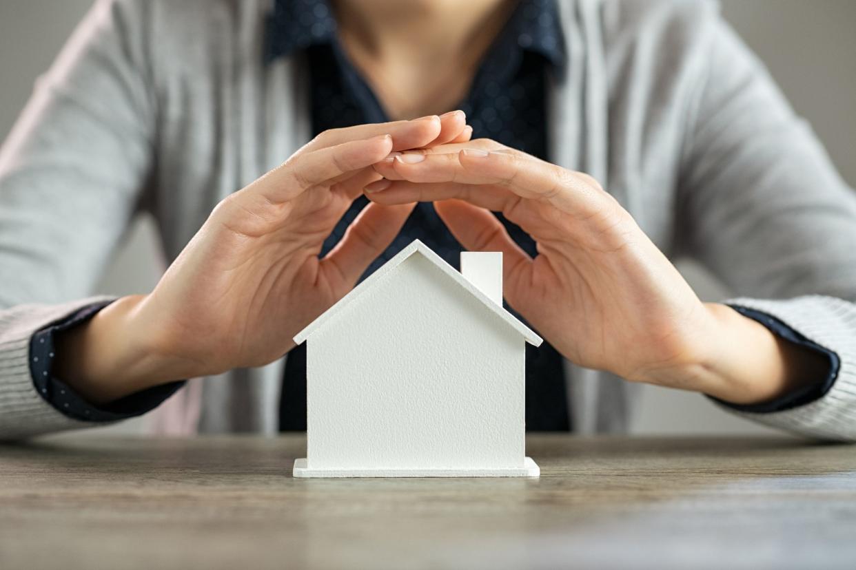 Close up of young woman hands protecting white model of house on table. Detail of woman holding hands over cardboard figure of house at desk. Woman protecting her real estate investment on scale model of her home.