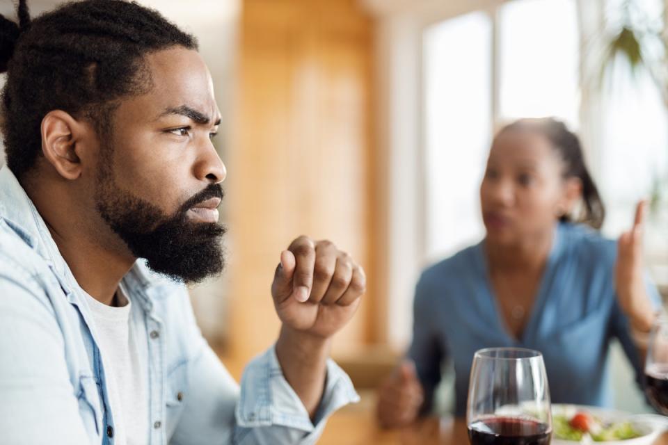 A bearded man looks into space while a blurry woman at the same table speaks to him.