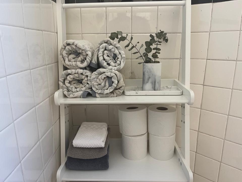 White shelf with four rolled towels, eucalyptus, gray and black folded towels, and four rolls of toilet paper