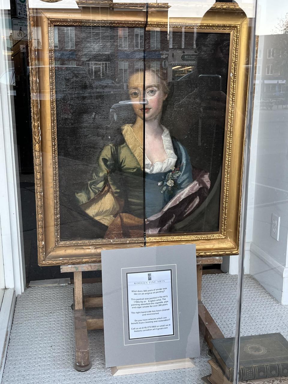 A framed painting of a woman in historical attire is displayed in a shop window. A placard with text is below the painting