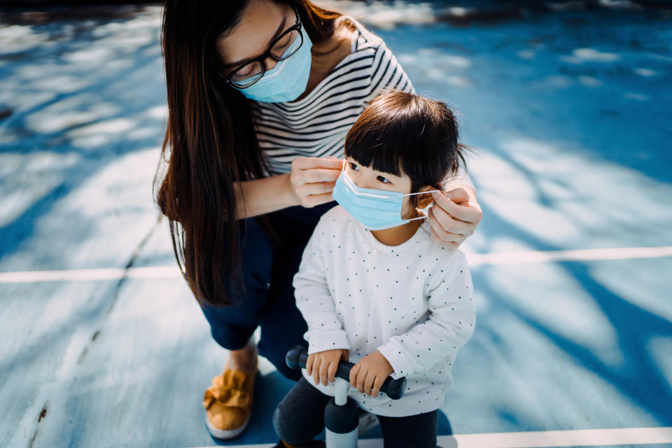 Young mother putting on surgical mask for little daughter in the playground to prevent the spread of cold and flu and viruses