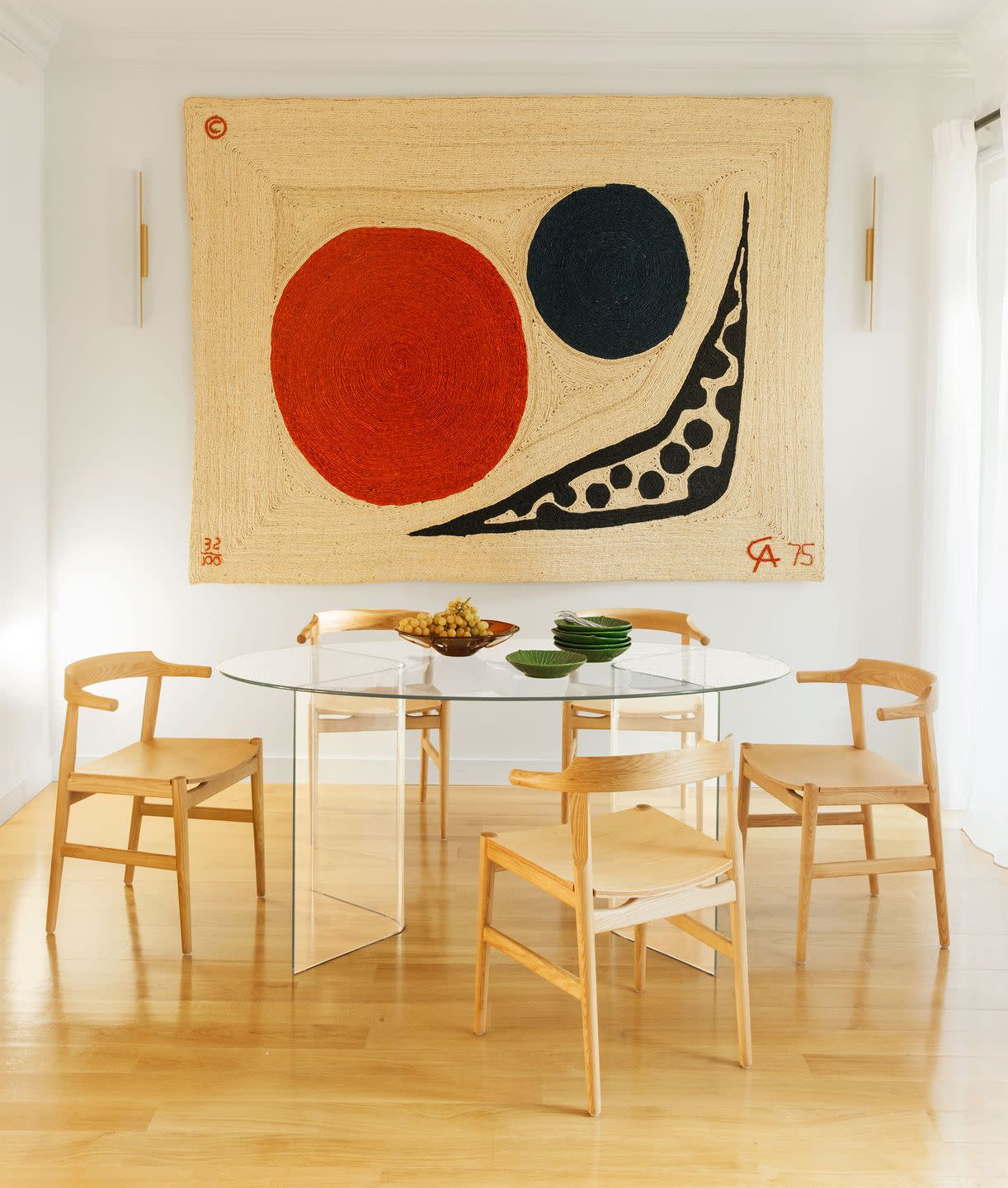 five simple wooden chairs surround a round glass dining table, the floor is wood, and on the wall is a large jute alexander calder tapestry with a red moon, a smaller black planet, and a boomerang shape