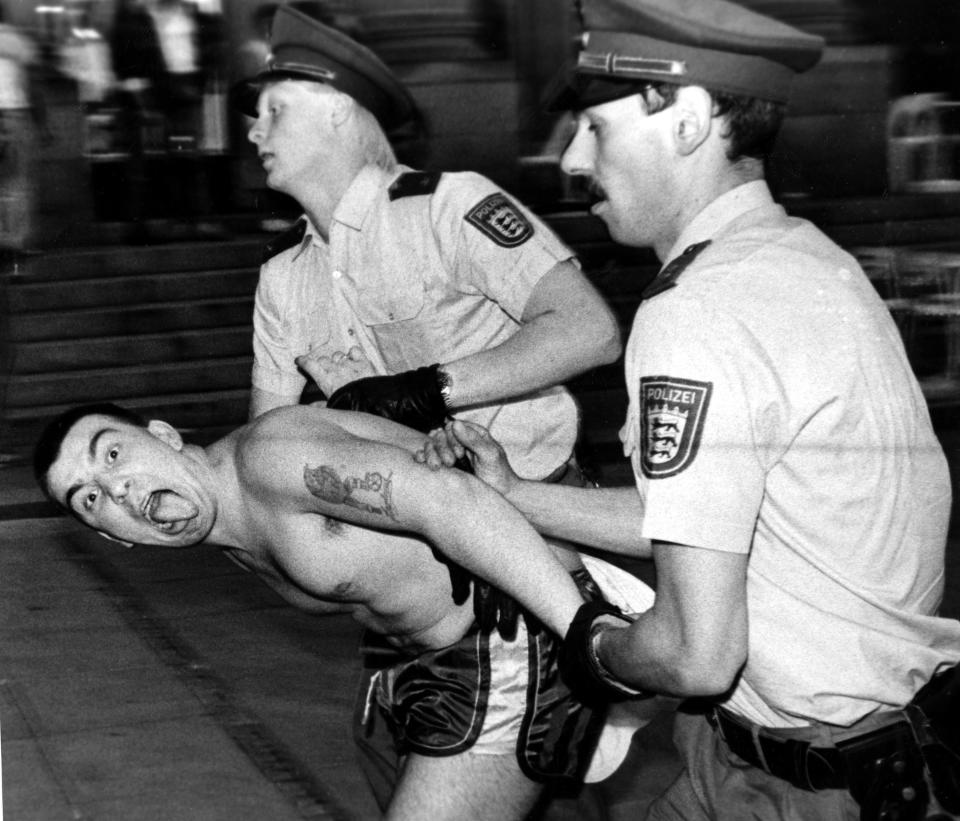 A shirtless man sticks out his tongue as he's restrained by two police officers.