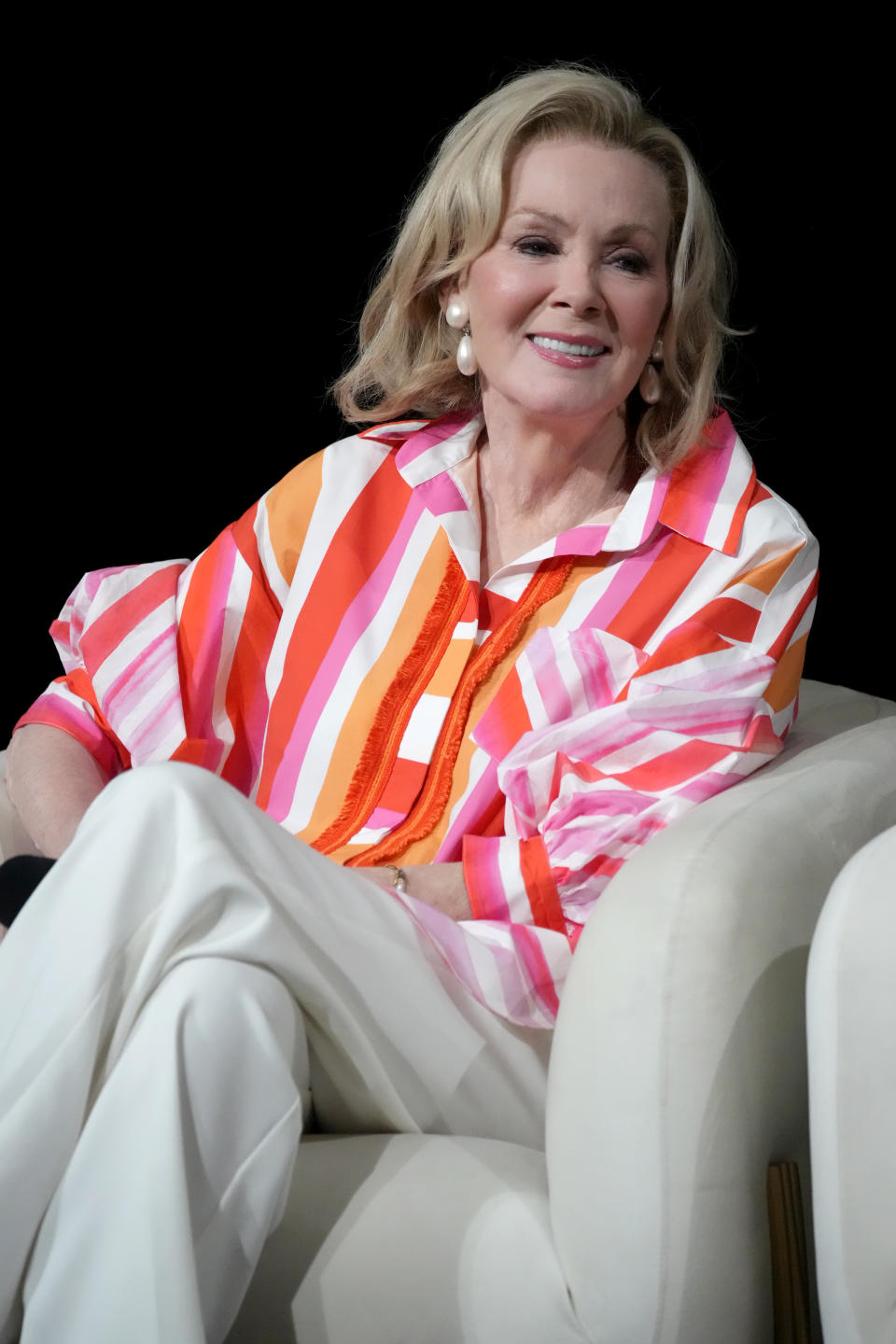 Jean Smart in a colorful striped blouse and white pants, sitting on a chair at a public event, smiling warmly