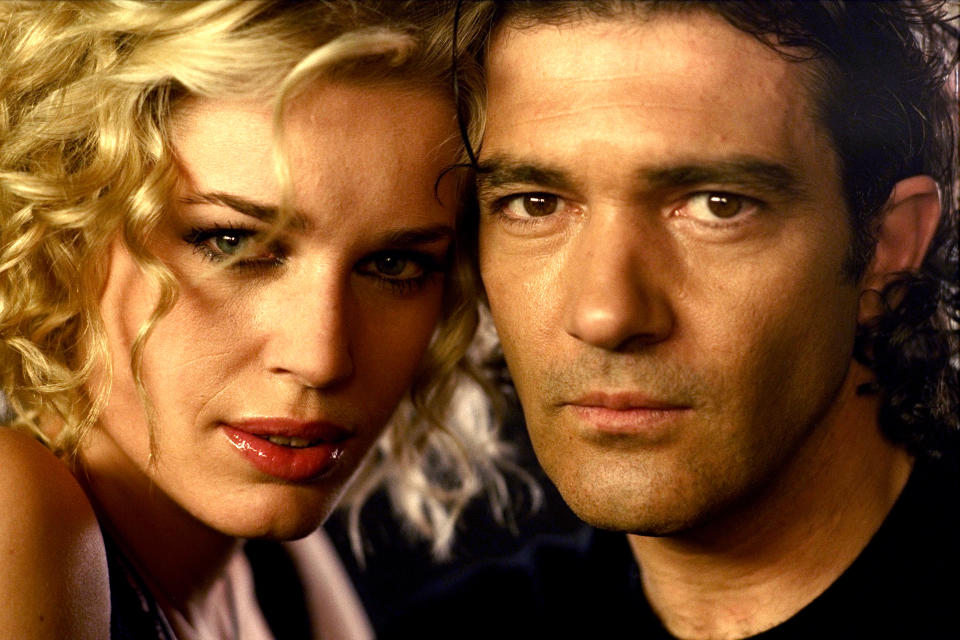 Rebecca Romijn and Antonio Banderas are closely posing, looking directly at the camera. Romijn has curly hair and glossy lips; Banderas has a serious expression