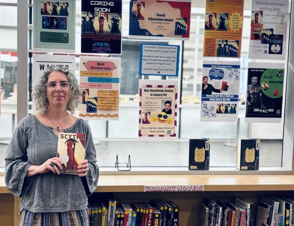 Sanford High School librarian Jennifer Raymond holds a copy of "Scythe," a Young Adult novel by bestselling author Neal Shusterman, who will visit SHS and the local middle school on April 4, 2024. Behind Raymond are posters students made to promote Shusterman's visit.