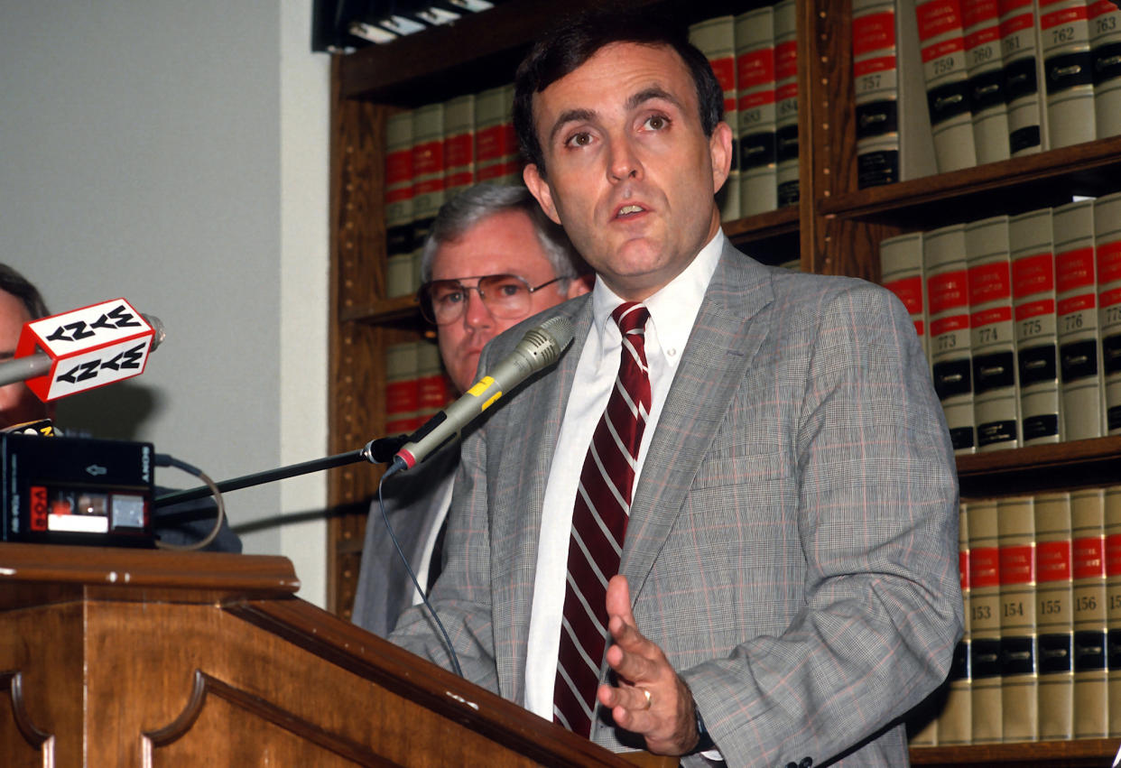 A young Rudy Giuliani at the microphone in front of a library of law volumes.