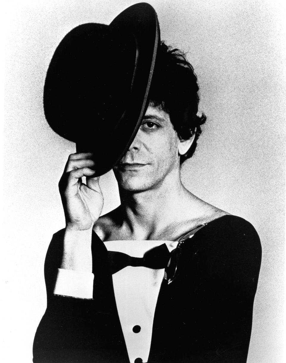 lou reed holds a top hat over one eye and part of his face, he looks straight ahead and wears a tuxedo top