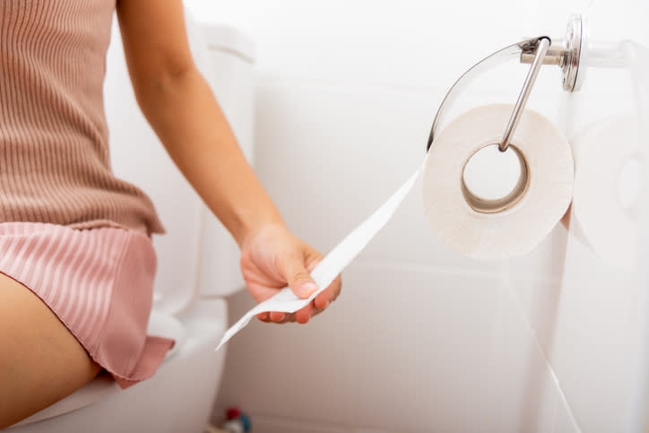 A person is sitting on a toilet, wearing a ribbed shirt and a skirt, holding a piece of toilet paper from a dispenser