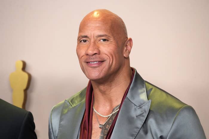 Dwayne Johnson wearing a satin suit, smiling at an event with a statuette in the background