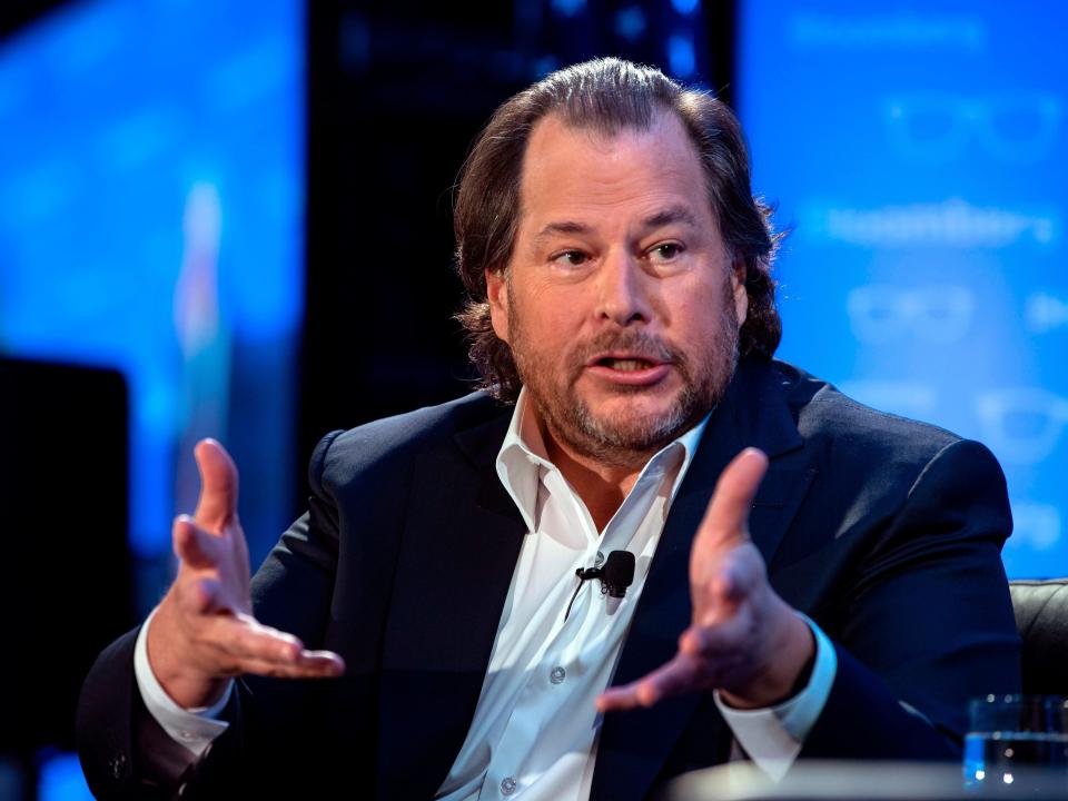 Le fondateur de Salesforce, Marc Benioff, vêtu d'une veste de sport et devant un fond bleu sur une scène, tend les mains vers l'avant lors d'une conférence.