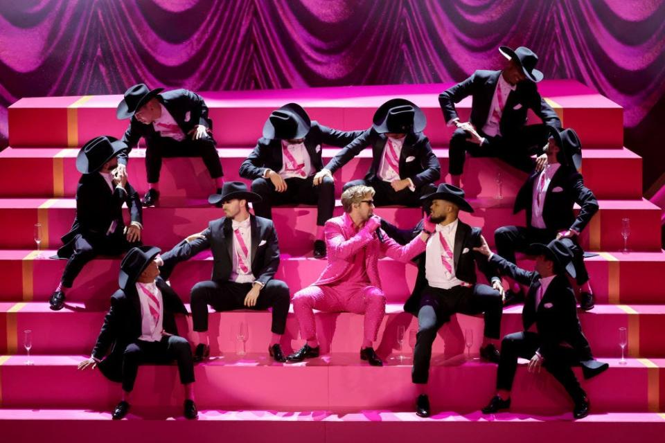 Ryan Gosling in a pink suit, sitting on pink steps and surrounded by male dancers in black and pink suits.