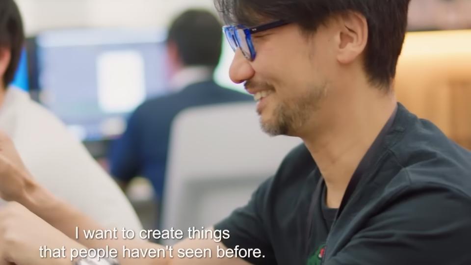 Still from the trailer to the documentary Hideo Kojima: Connecting Worlds. The game designer sits, smiling, in a work setting. A caption reads, 