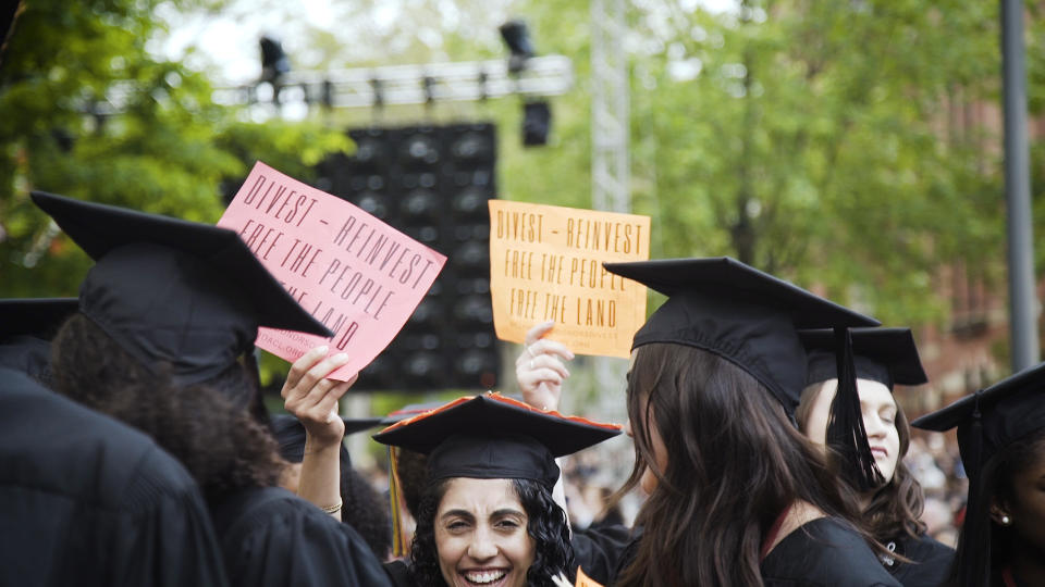 In a show of protest, Harvard graduates hold up red and orange signs that say, 