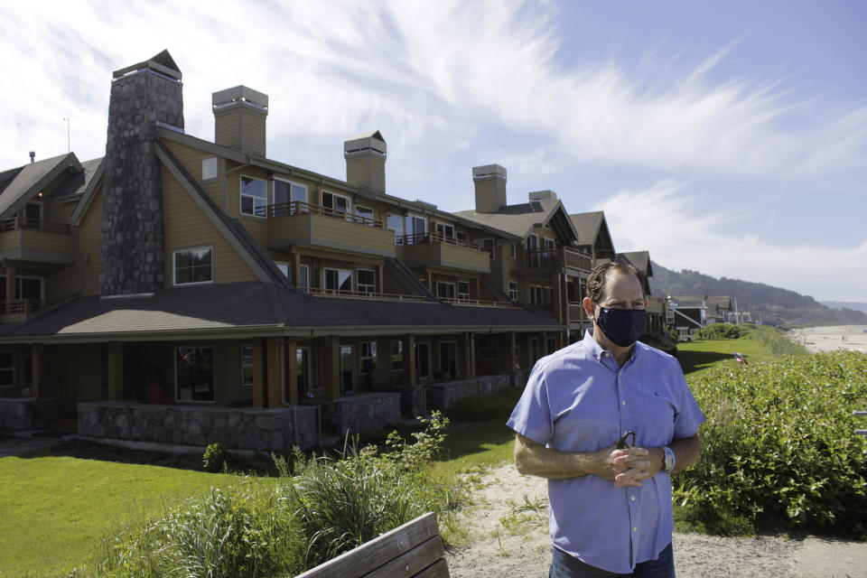 In this Thursday, May 28, 2020, photo, Patrick Nofield, whose company, Escape Lodging, owns and operates four hotels in Cannon Beach, Ore., stands in front of the flagship property, The Ocean Lodge, while wearing a face mask due to the novel coronavirus. With summer looming, Cannon Beach and thousands of other small, tourist-dependent towns nationwide are struggling to balance fears of contagion with their economic survival in what could be a make-or-break summer. (AP Photo/Gillian Flaccus)