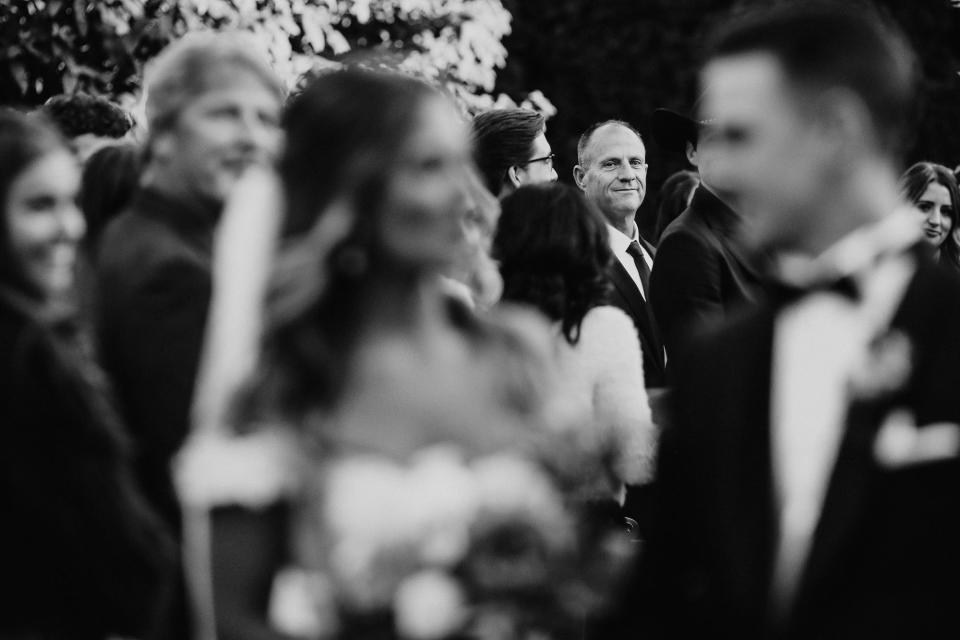 A father looks at a bride and groom as they say their vows in black and white.