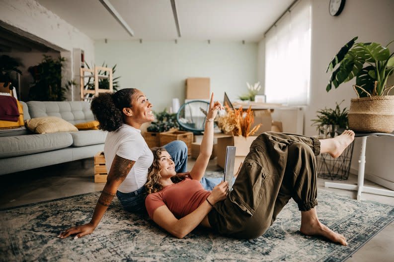 A couple smiles and looks at a tablet while sitting on the floor in their apartment.