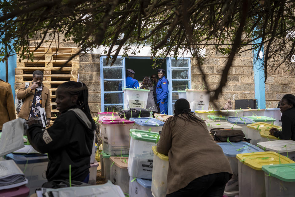 A policeman looks out as electoral workers stand with ballot boxes lined up and ready to be stored at a collection and tallying center in Nairobi, Kenya Wednesday, Aug. 10, 2022. Kenyans are waiting for the results of a close but calm presidential election in which the turnout was lower than usual. (AP Photo/Ben Curtis)