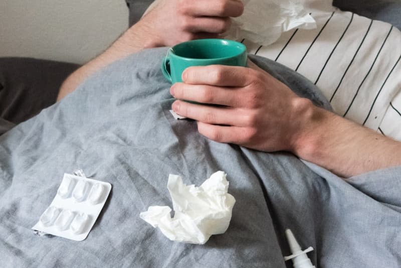 A man lies in bed with handkerchiefs, teacup, nasal spray and tablets. A wave of flu infections in Germany has continued to grow and now affects all age groups, according to a weekly update from Germany's public health authority. Philip Dulian/dpa