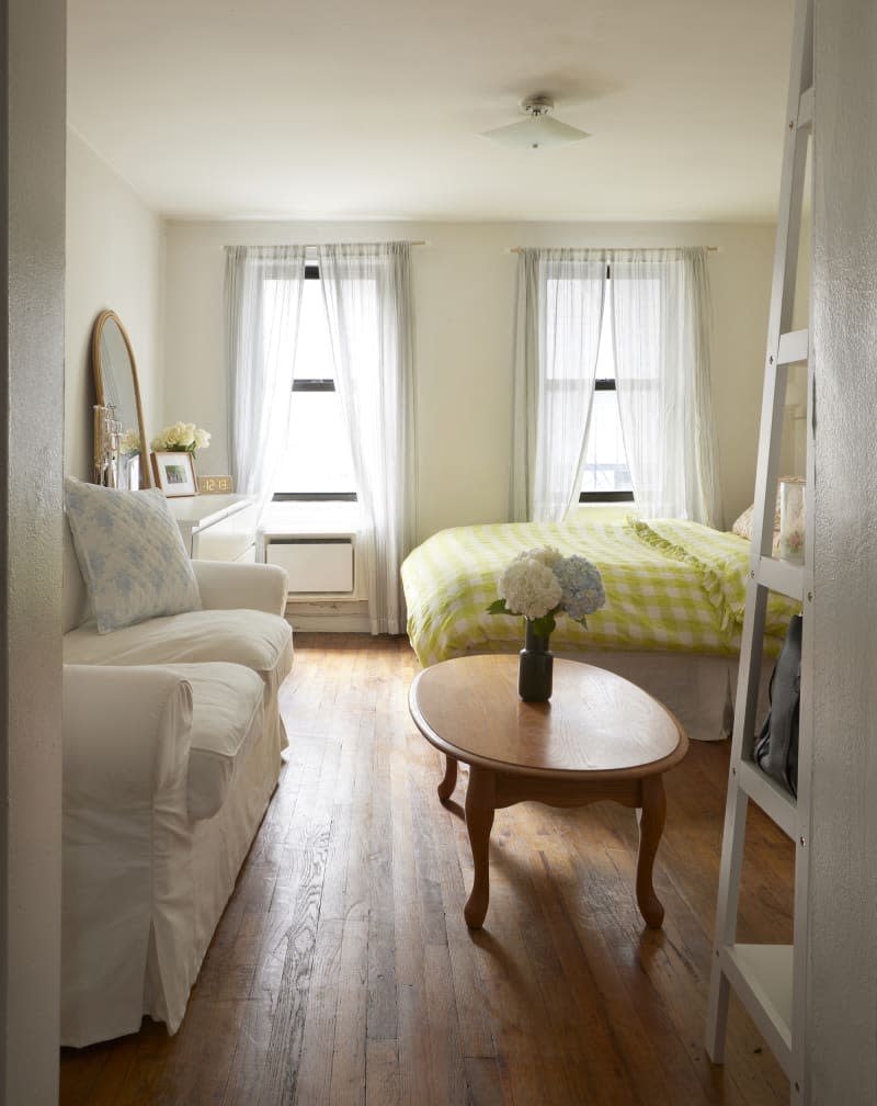 Living space with bed with chartreuse and white gingham duvet with floral and plaid pillowcases, straw round bedside table with case of yellow flowers, wood shoe rack with wall mount TV over it, windows with sheer white curtains, wood floor, White sofa with light blue and white pillows, oval wood table with vase of white and light blue hydrangeas