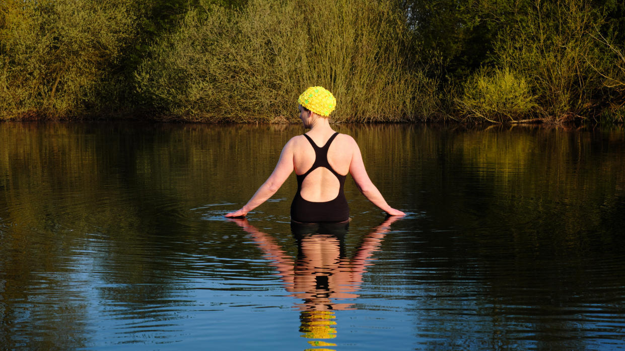  UK, Essex, female wild swimmer standing in a lake touching the water at sunrise. 