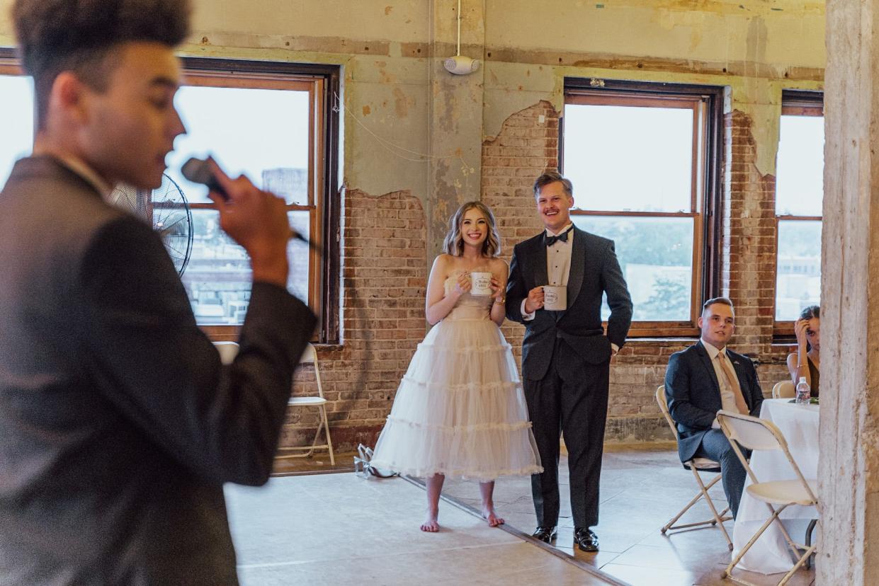 A bride and groom watch someone give a toast at their wedding reception.