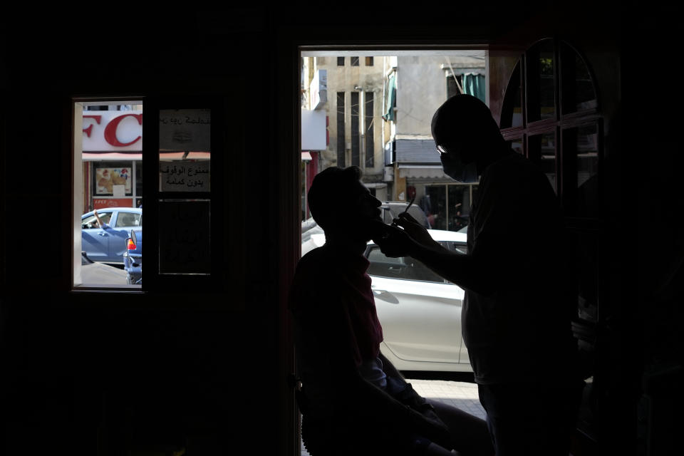 A Lebanese barber shaves the beard of a customer at the door of his shop during a power outage in a southern suburb of Beirut, Lebanon, Wednesday, Aug. 11, 2021. Lebanon, which is mired in multiple crises including a devastating economic crisis, has faced months of severe fuel shortages that have prompted long lines at gas stations and plunged the small country, dependent on private generators for power, into long hours of darkness. (AP Photo/Hassan Ammar)
