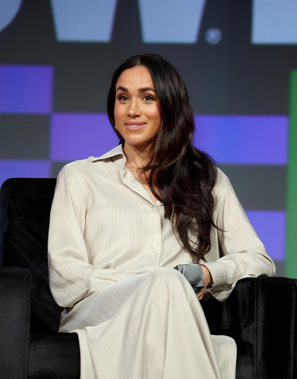 Meghan Markle at a public speaking event, sitting on a black chair, wearing a light-colored long-sleeved dress, and holding a microphone