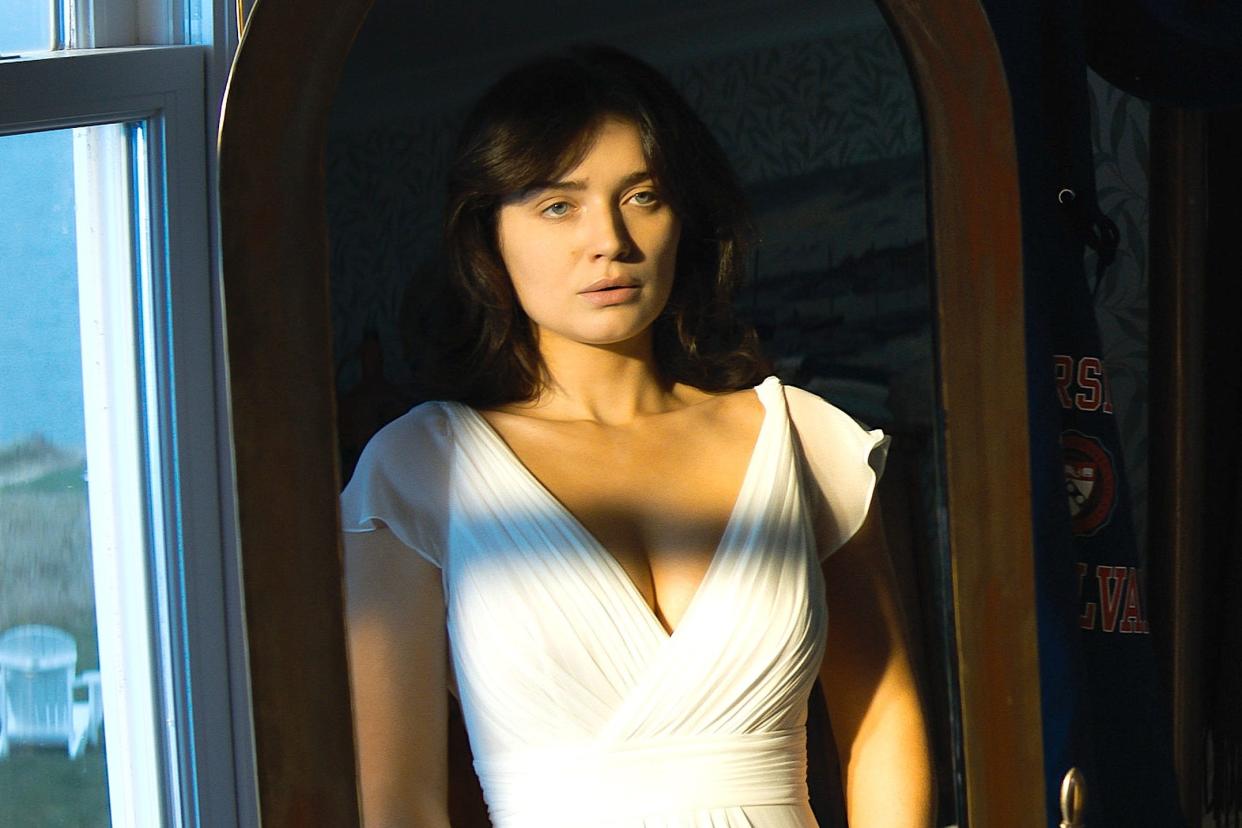 A woman in a white wedding dress looking at herself in a wooden mirror in a bedroom.