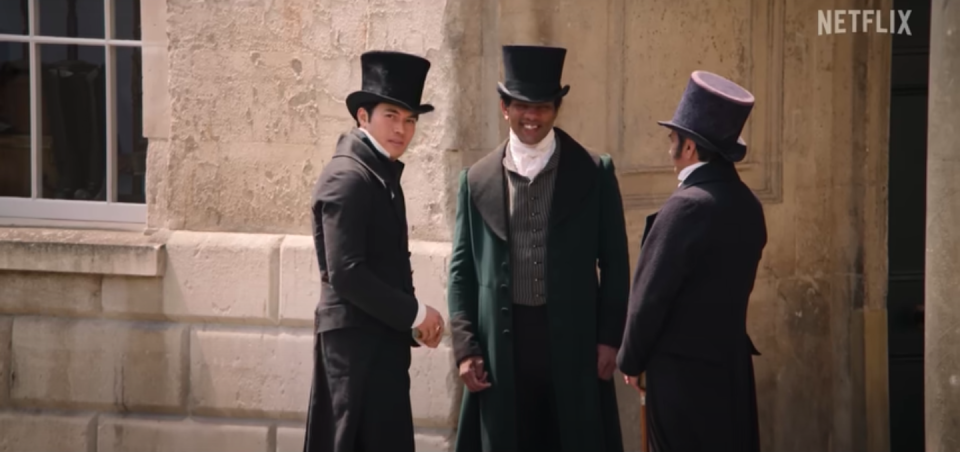 Henry Golding wearing a top hat while standing next to to other gentlemen