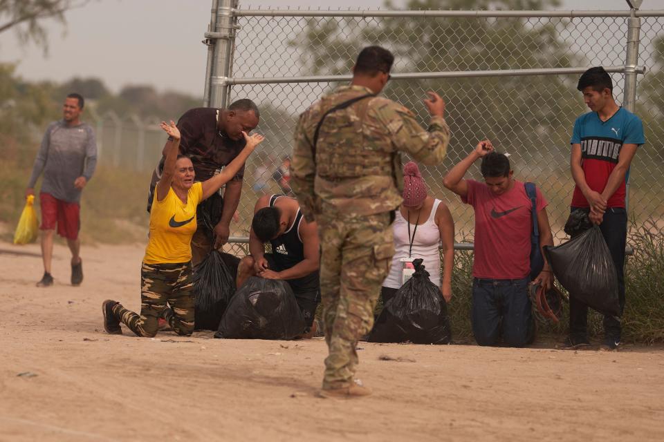 Half a dozen migrants kneel or stand by a cyclone fence as they await processing by the Border Patrol.