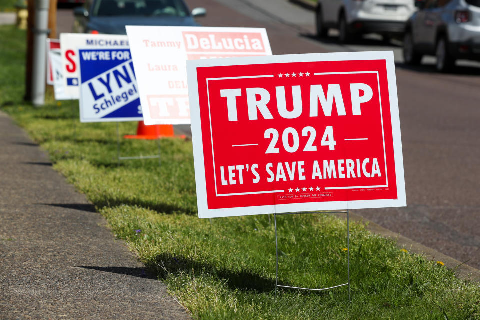 Several political campaign signs, including a prominent "Trump 2024: Let's Save America" sign, are displayed along a grassy roadside