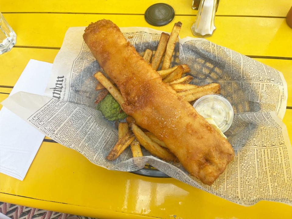 A large piece of fried fish over a bed of french fries on a newspaper-lined bowl on a yellow table