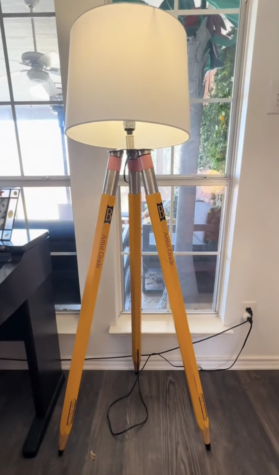 A unique lamp with a shade supported by three oversized pencils as its base, set in a room by a window