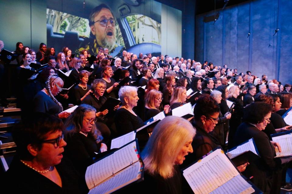 Performers wearing black look at their music books and sing as a group.