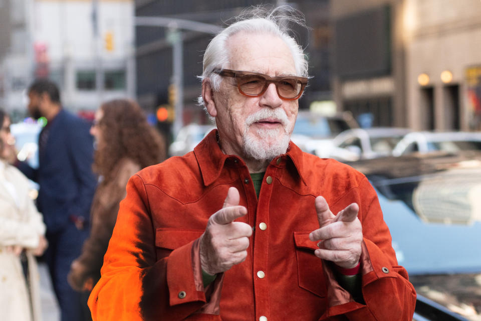 Brian Cox in an jacket pointing towards the camera while on a street