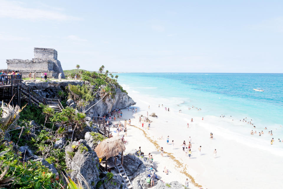 Beach with visitors below ancient ruins, clear sky, and a traditional hut on the left