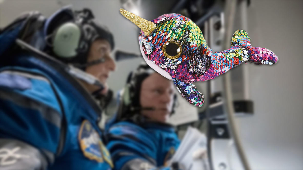  A plush, colorful toy narwhal floats in zero gravity with two astronauts in blue flight suits in the background. 