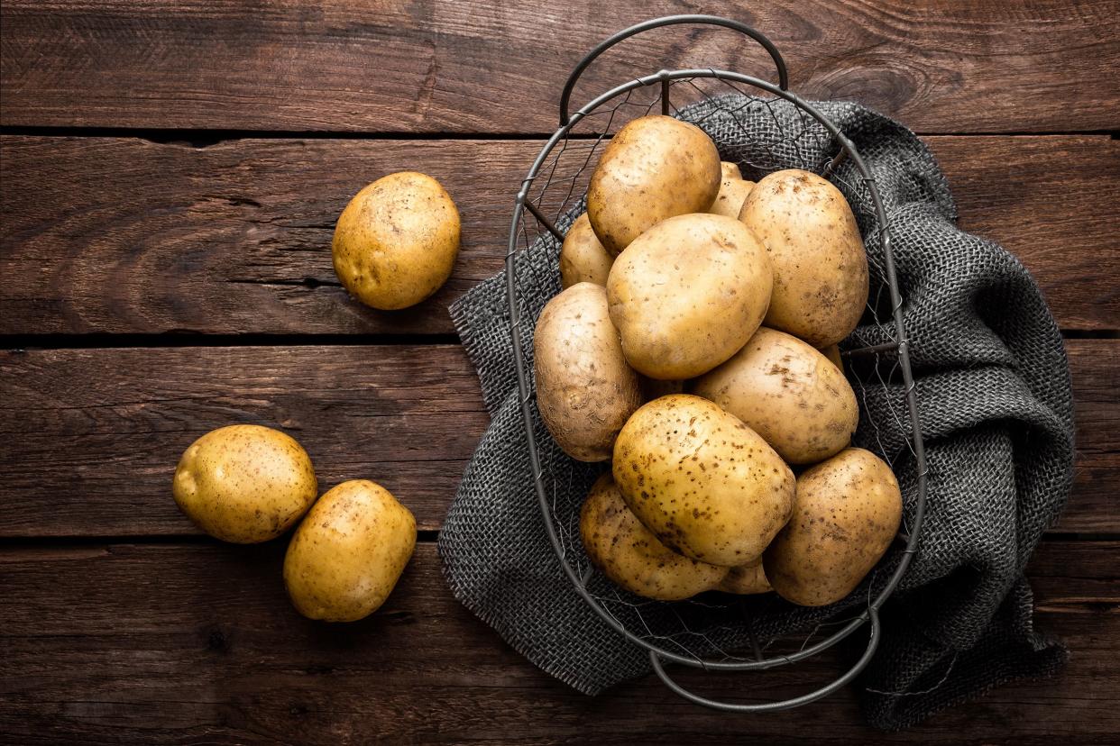 A pile of russet potatoes in a wire basket on a dark grey napkin on a rustic wooden table with three russet potatoes scattered