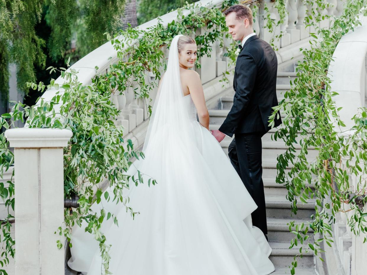 A bride and groom walk up a staircase covered in greenery and look over their shoulders.